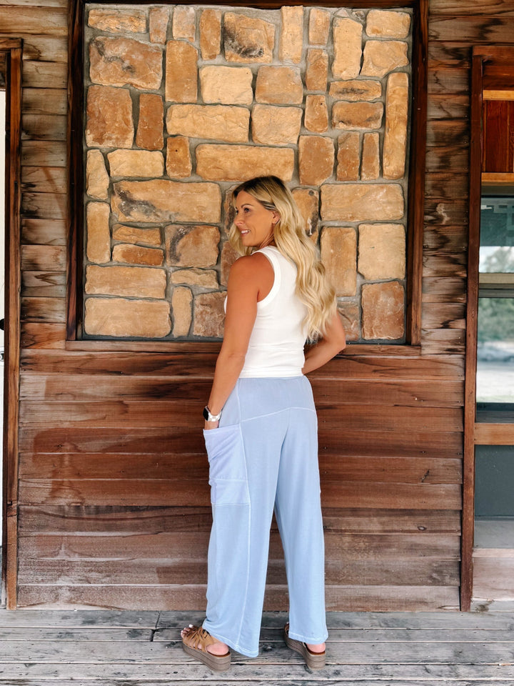 A woman stands on a rustic wooden porch in front of a stone wall, wearing light blue wide-leg cargo pants, a white tank top, and brown sandals.