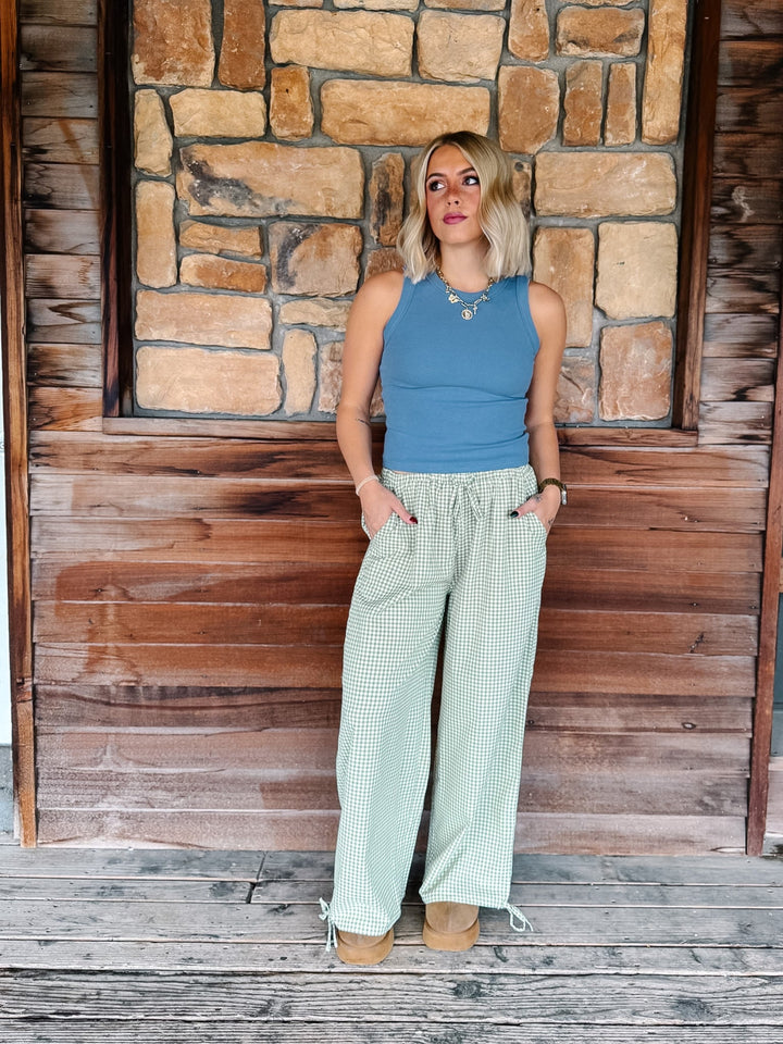 A woman stands on a rustic wooden porch in front of a stone wall, wearing a sleeveless blue top and light sage green wide-leg checkered pants.