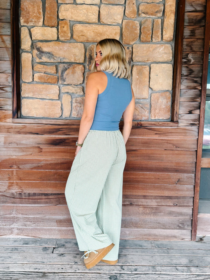 A woman stands on a wooden porch in front of a stone wall, wearing a sleeveless denim blue top and light sage green wide-leg plaid pants with tan platform sneakers.