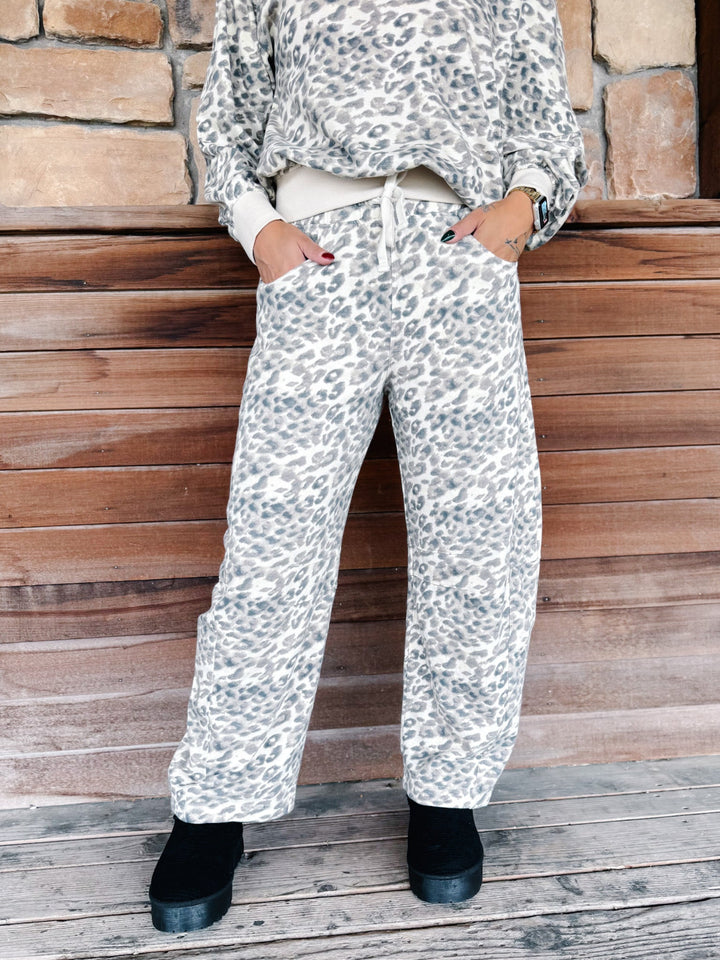 A person wearing loose-fit leopard-print pants with a white drawstring waist, paired with a matching top and black boots, standing against a rustic wood and stone wall.