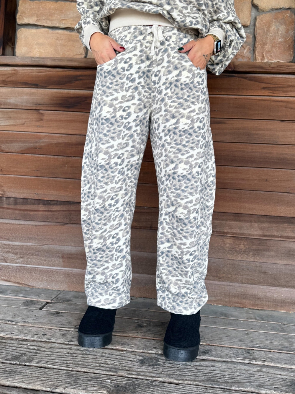 A person wearing loose, wide-leg sweatpants with an allover gray and black leopard print on a light beige base, paired with a matching top and black boots, standing against a rustic wooden wall.