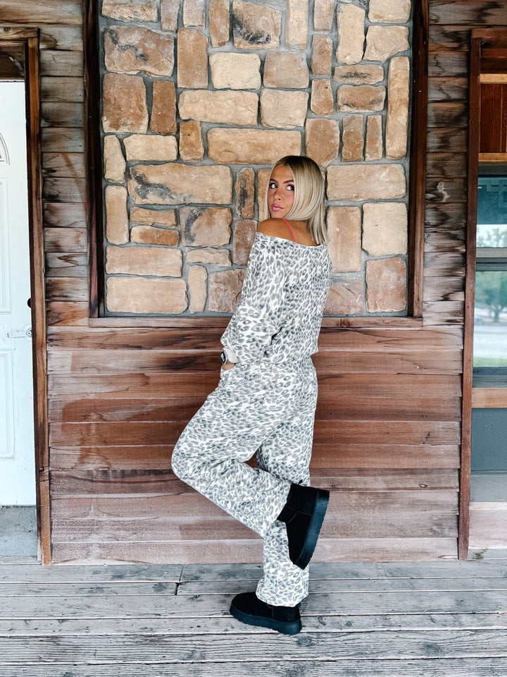 A woman wearing a matching off-the-shoulder long-sleeve top and wide-leg pants set in a black and white abstract pattern, posing on a rustic wooden porch with a stone wall background.