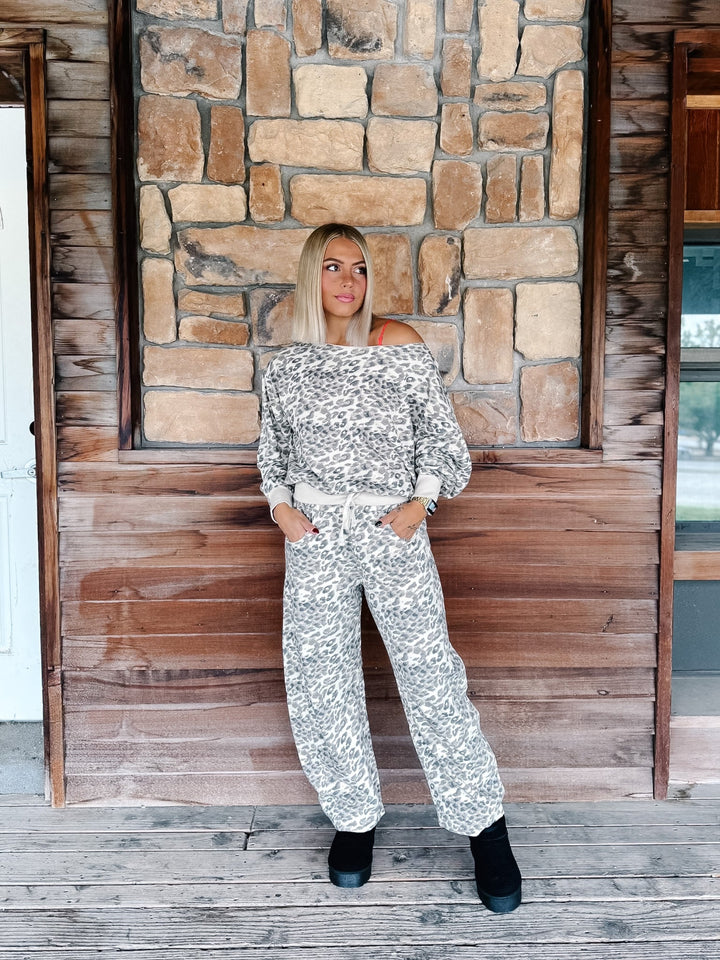 A woman modeling a matching off-the-shoulder long-sleeve top and wide-leg pants set in a gray and white floral pattern, standing on a rustic wooden porch in front of a stone wall.