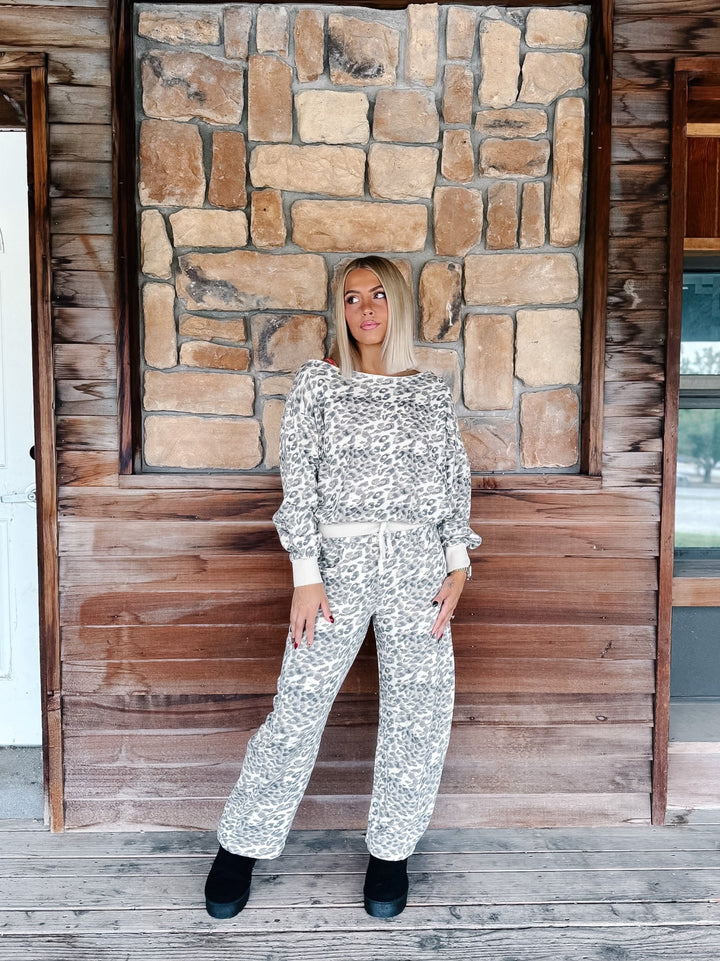 A woman modeling a matching two-piece loungewear set featuring a white and gray leopard print pattern, standing in front of a rustic stone and wood wall.