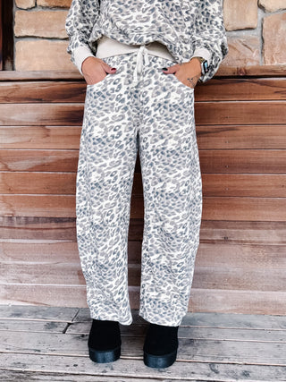 A person wearing loose, wide-leg pants with an allover leopard print in gray and white, paired with a matching top and black boots, standing against a rustic wooden wall.