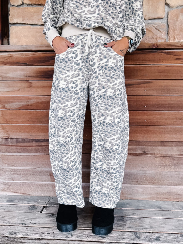 A person wearing loose, wide-leg pants with an allover leopard print in gray and white, paired with a matching top and black boots, standing against a rustic wooden wall.