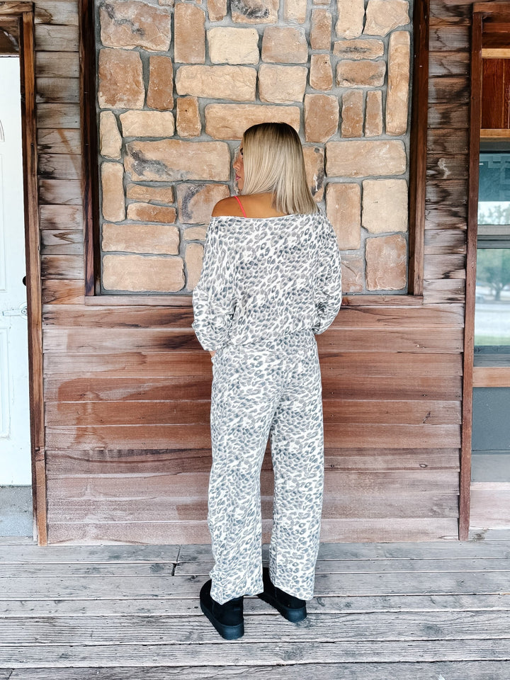 A woman stands on a rustic wooden porch facing a stone wall, wearing a white off-the-shoulder jumpsuit with a gray and black animal print pattern.