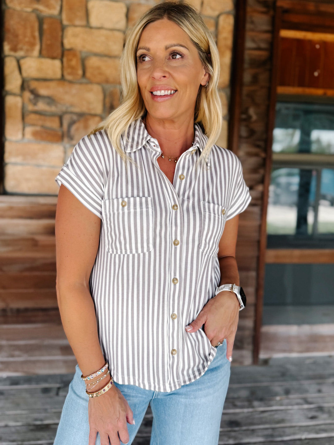 A woman stands on a wooden porch in front of a stone and wood wall, wearing a white short-sleeve button-up shirt with thin vertical mocha stripes