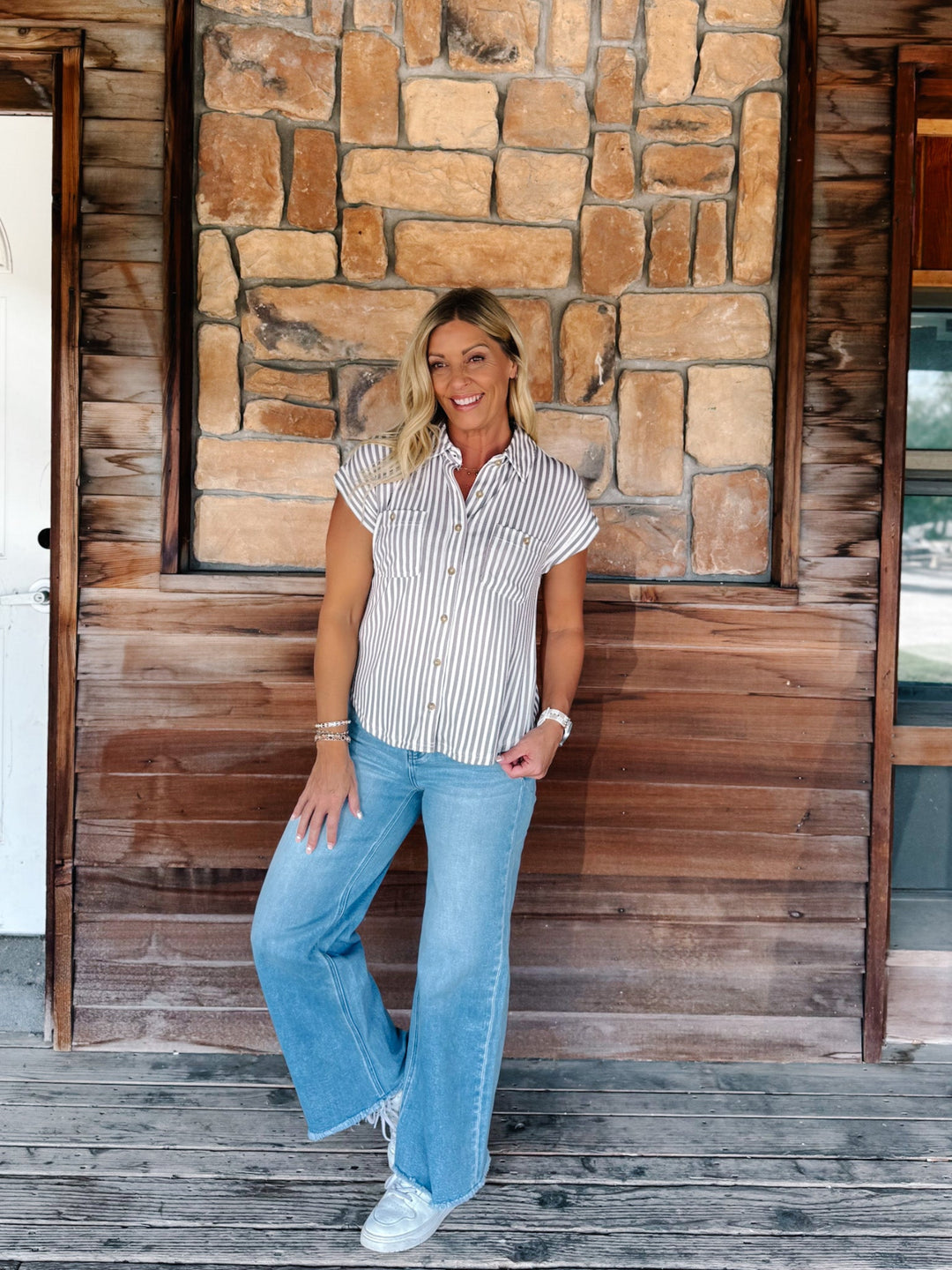 A woman stands on a wooden porch in front of a stone and wood wall, wearing a white short-sleeve button-up shirt with thin vertical mocha stripes, light blue wide-leg jeans, and white sneakers.