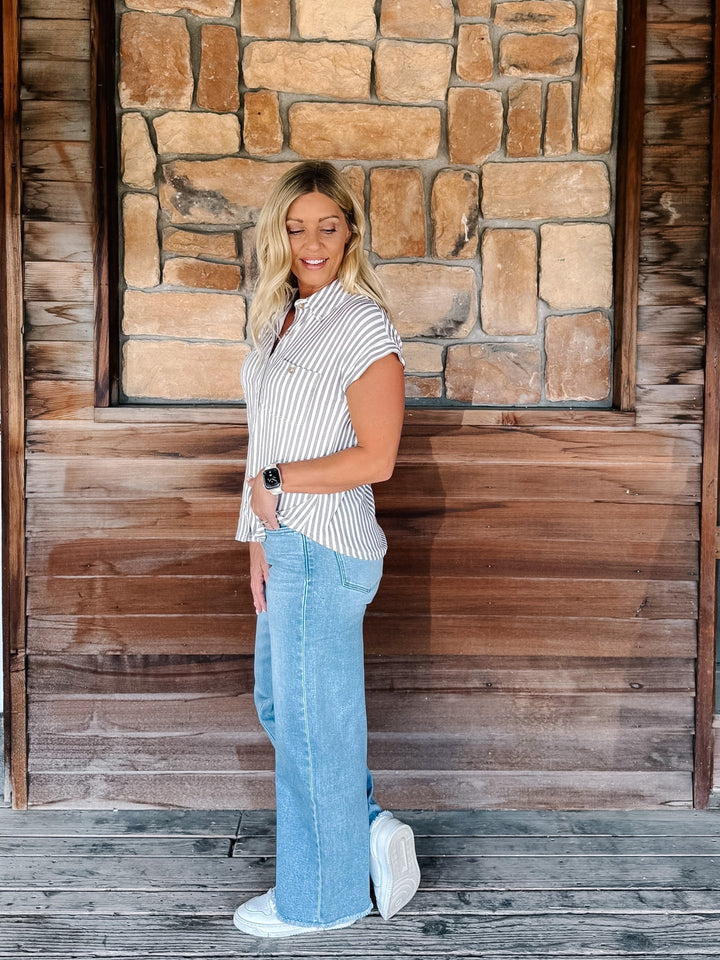 A woman stands on a wooden porch in front of a stone and wood wall, wearing a white short-sleeve button-up shirt with thin vertical mocha stripes, light blue wide-leg jeans, and white sneakers.