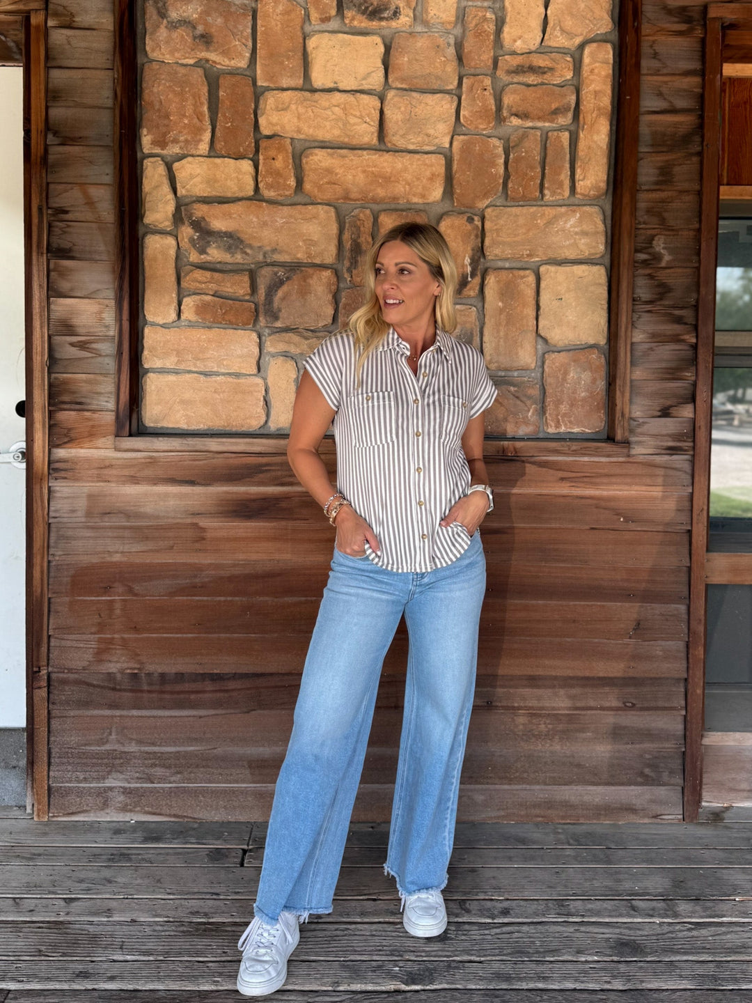 A woman stands on a wooden porch in front of a stone and wood wall, wearing a white short-sleeve button-up shirt with thin vertical mocha stripes, light blue wide-leg jeans, and white sneakers.