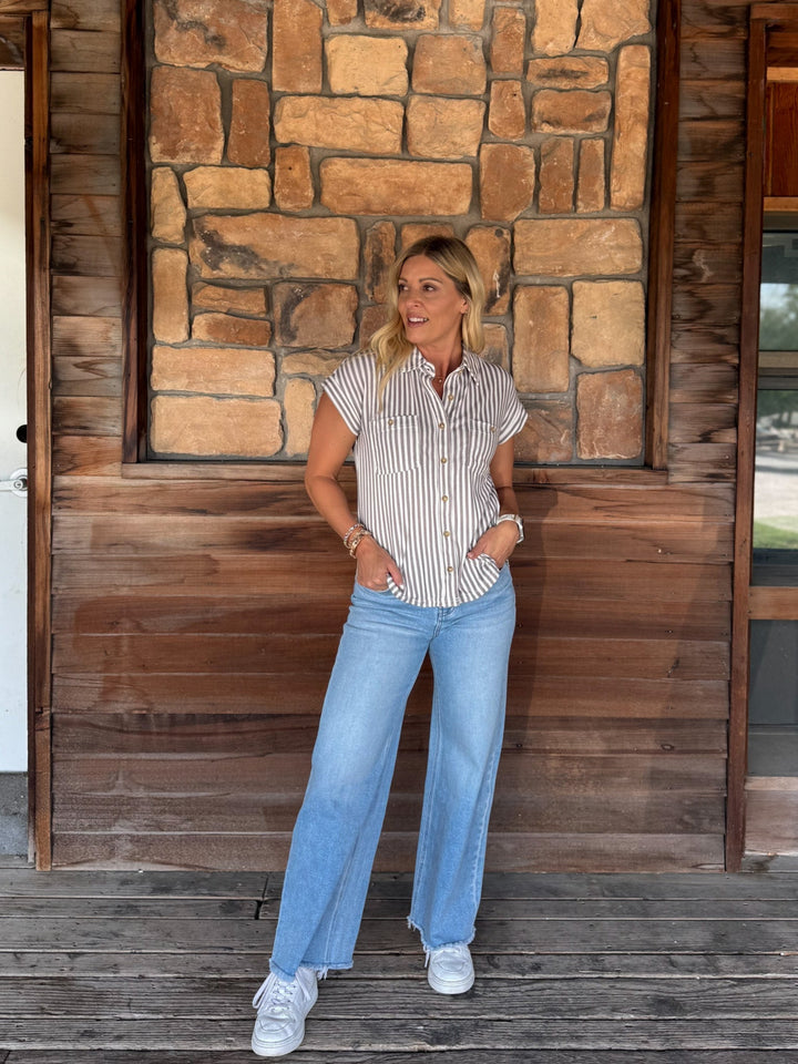 A woman stands on a wooden porch in front of a stone and wood wall, wearing a white short-sleeve button-up shirt with thin vertical mocha stripes, light blue wide-leg jeans, and white sneakers.