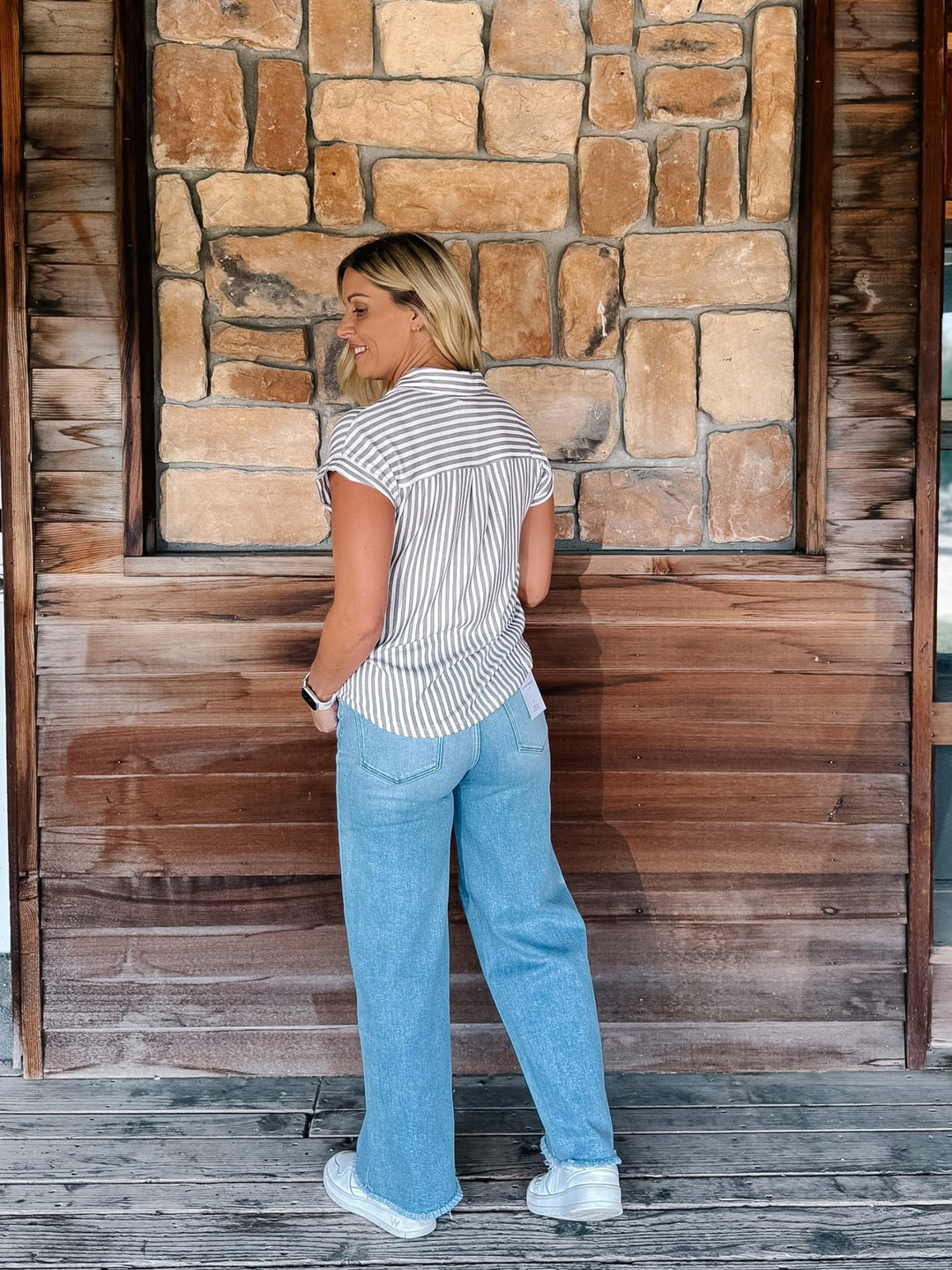 A woman stands on a wooden porch in front of a stone and wood wall, wearing a white short-sleeve button-up shirt with thin vertical mocha stripes, light blue wide-leg jeans, and white sneakers.
