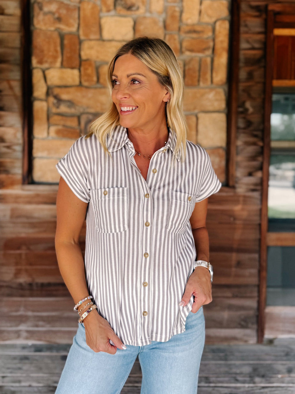 A woman wearing a short-sleeve button-up shirt with vertical mocha and white stripes, paired with light blue jeans, standing in front of a rustic stone and wood wall.