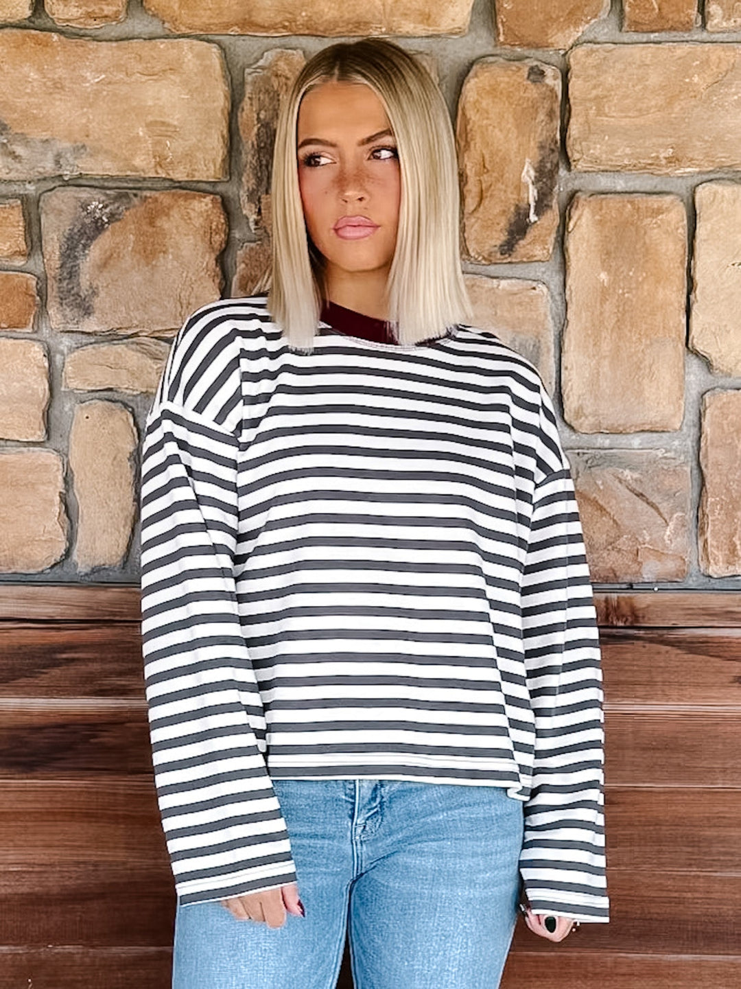 A woman with blonde hair wearing a charcoal and white horizontally striped long-sleeve top with maroon neckline detail and light blue jeans, standing in front of a rustic stone wall.