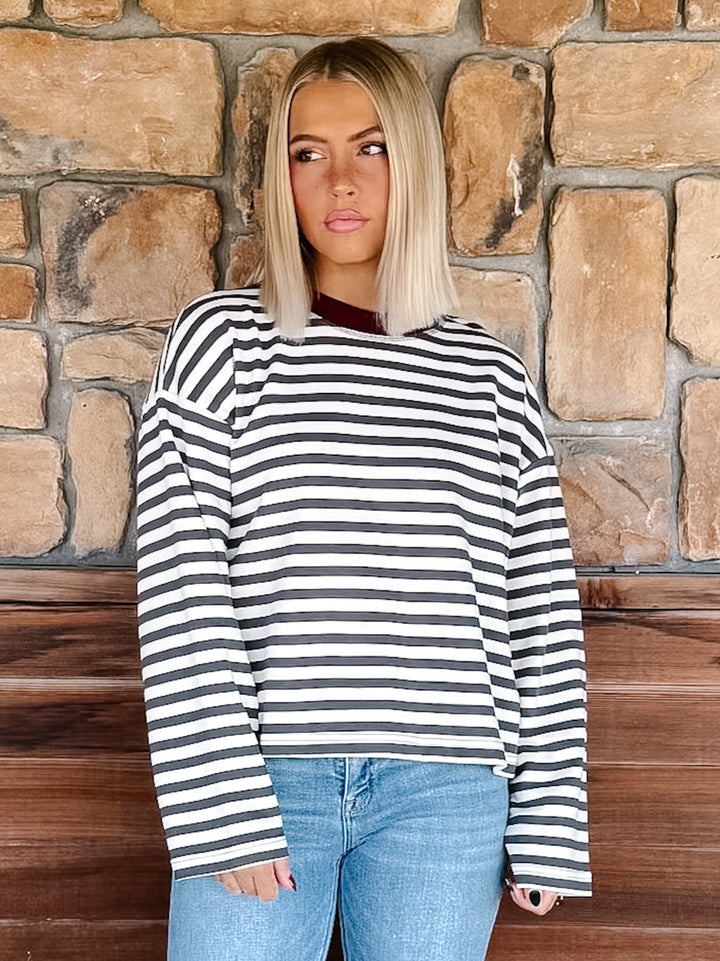 A woman with blonde hair wearing a charcoal and white horizontally striped long-sleeve top with maroon neckline detail and light blue jeans, standing in front of a rustic stone wall.