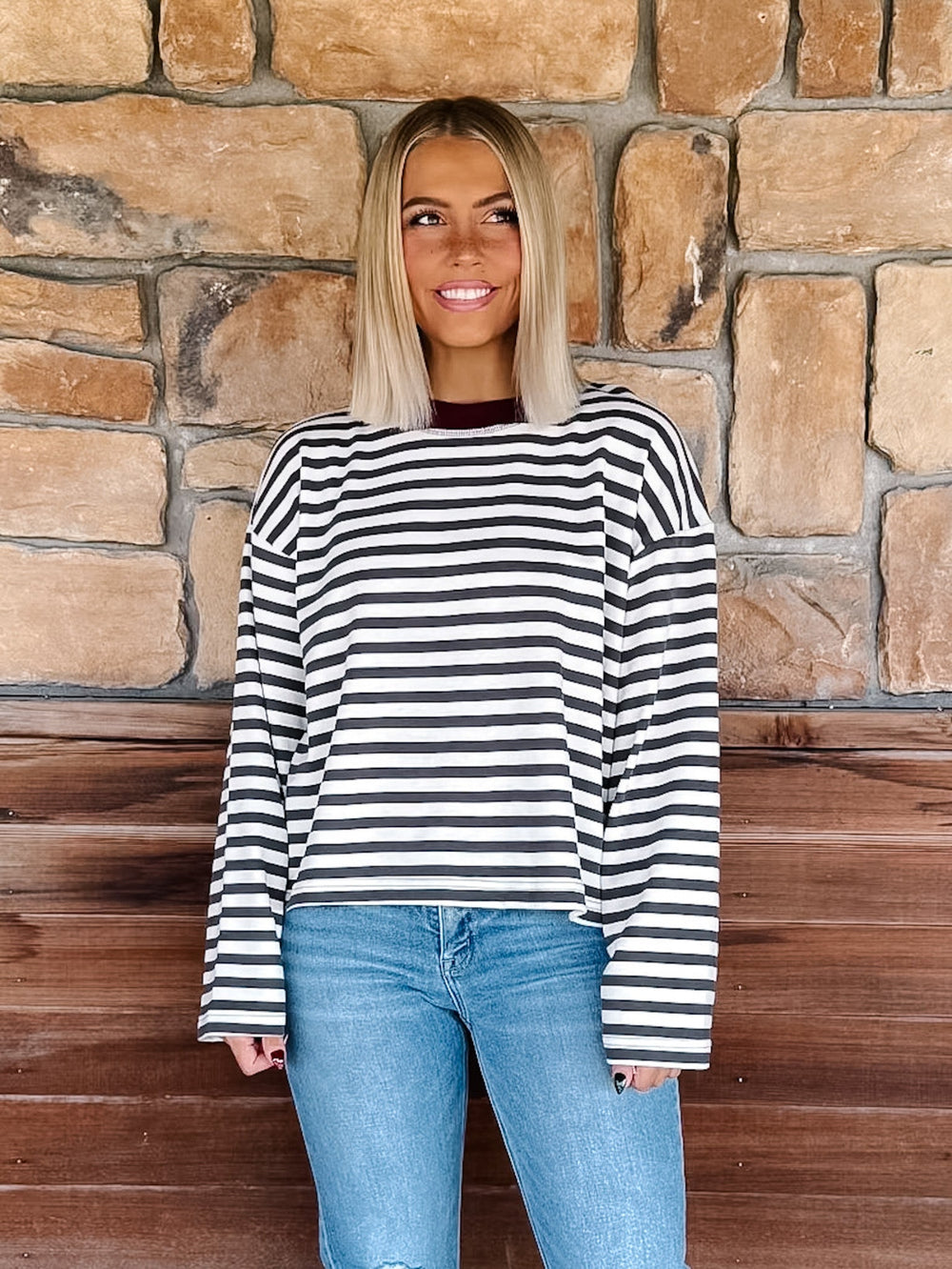 A woman with blonde hair wearing a charcoal and white horizontally striped long-sleeve top with maroon neckline detail and light blue jeans, standing in front of a rustic stone wall.