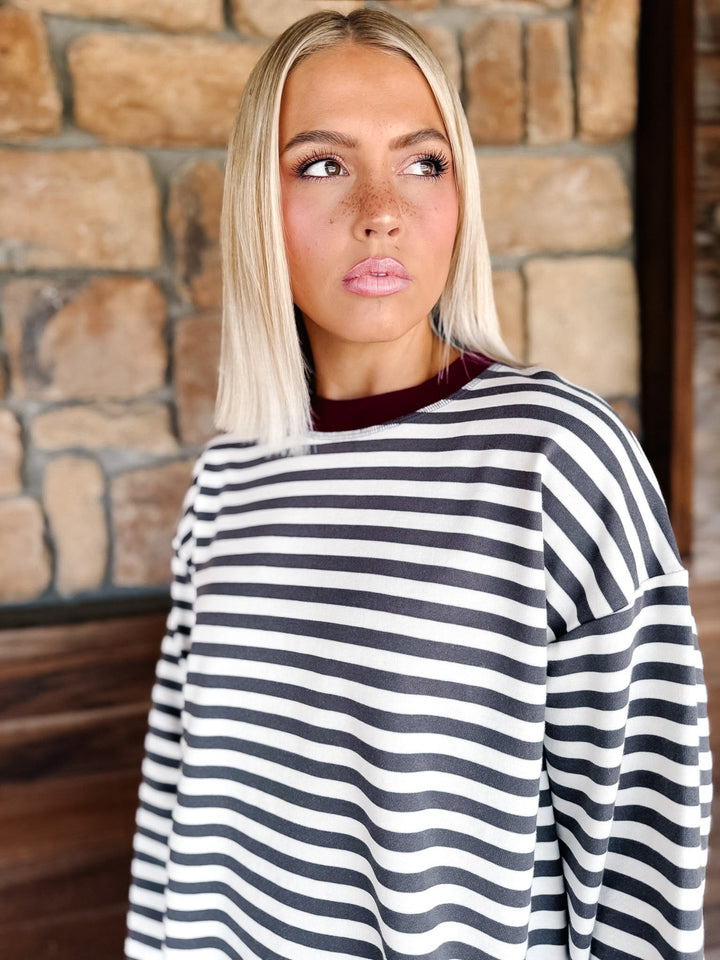 A woman with blonde hair wearing a charcoal and white horizontally striped long-sleeve top with maroon neckline detail and light blue jeans, standing in front of a rustic stone wall.