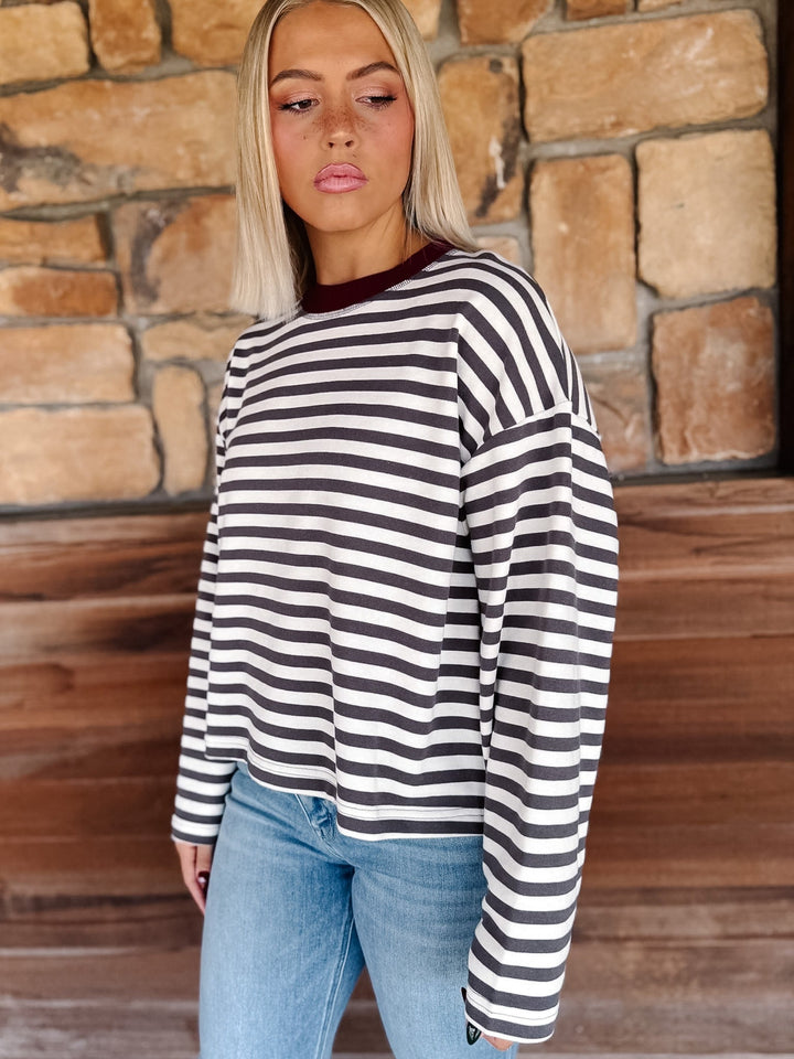 A woman with blonde hair wearing a charcoal and white horizontally striped long-sleeve top with maroon neckline detail and light blue jeans, standing in front of a rustic stone wall.