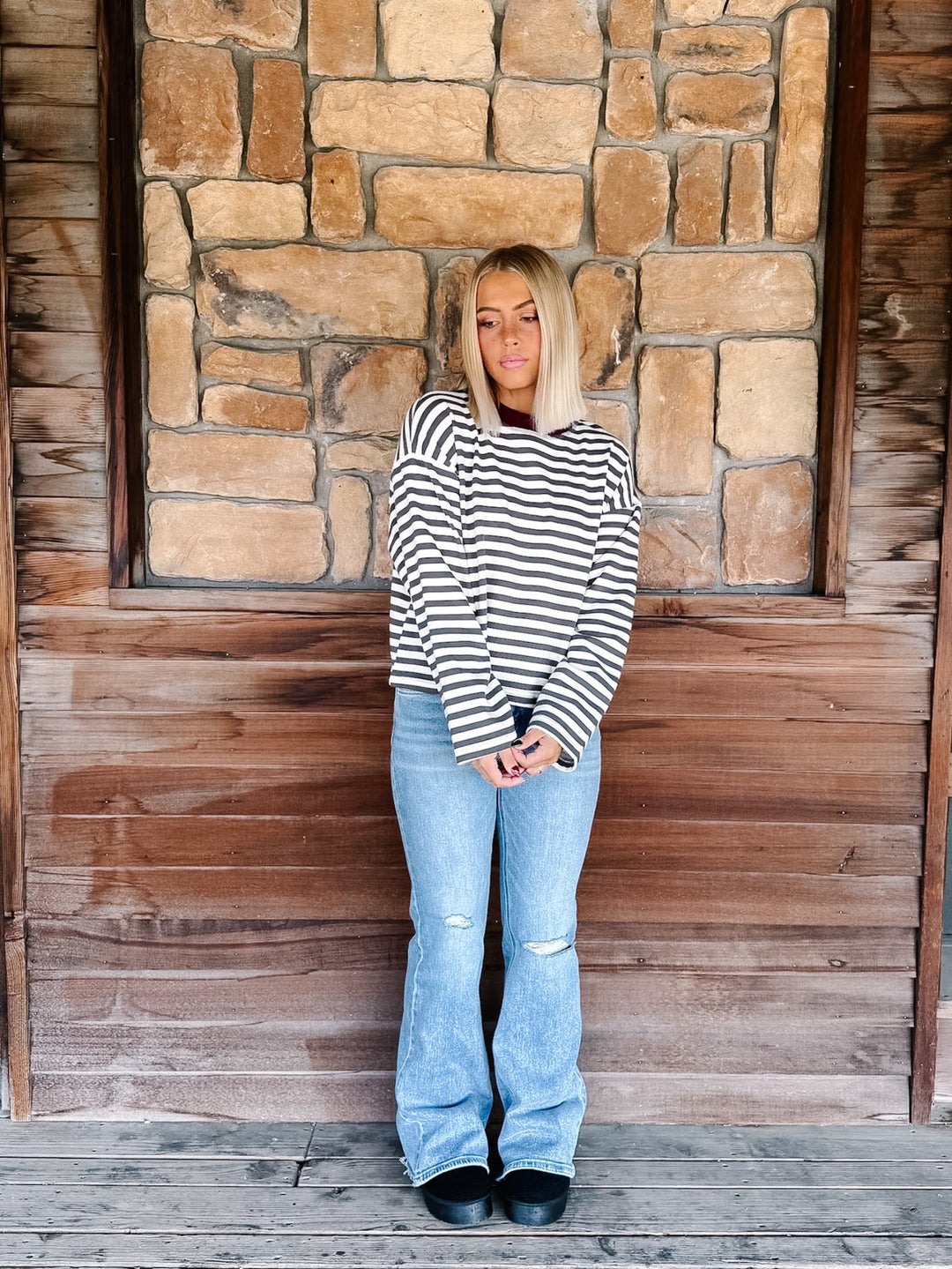 A woman with blonde hair wearing a charcoal and white horizontally striped long-sleeve top with maroon neckline detail and light blue jeans, standing in front of a rustic stone wall.