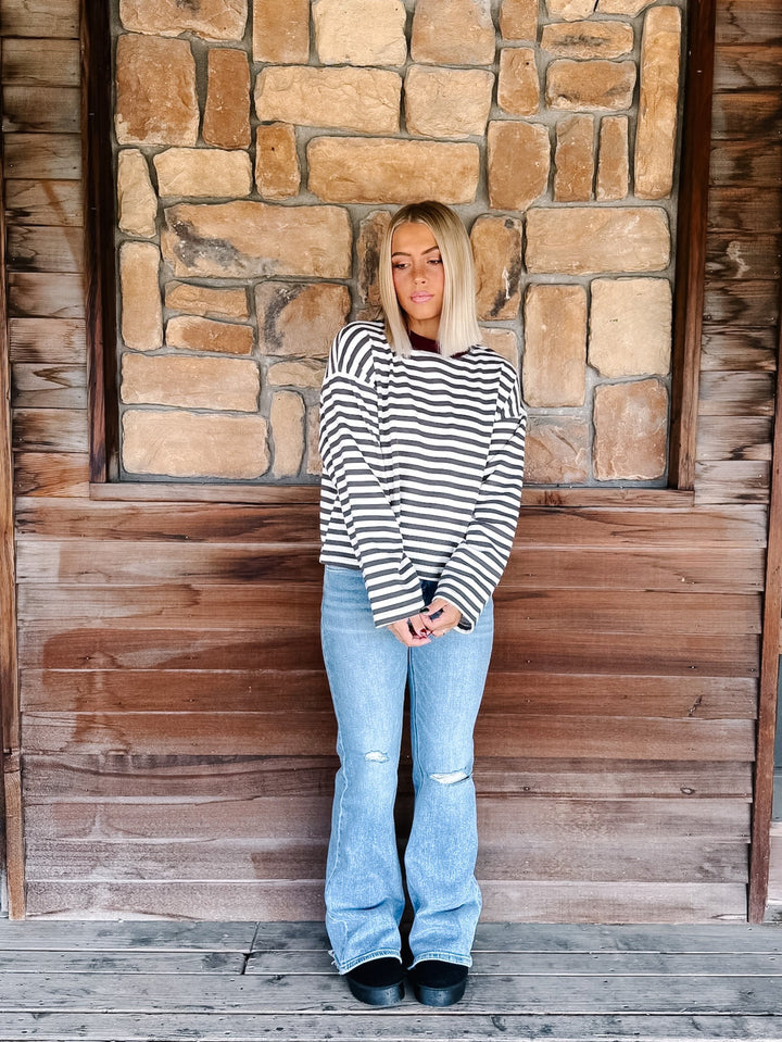 A woman with blonde hair wearing a charcoal and white horizontally striped long-sleeve top with maroon neckline detail and light blue jeans, standing in front of a rustic stone wall.