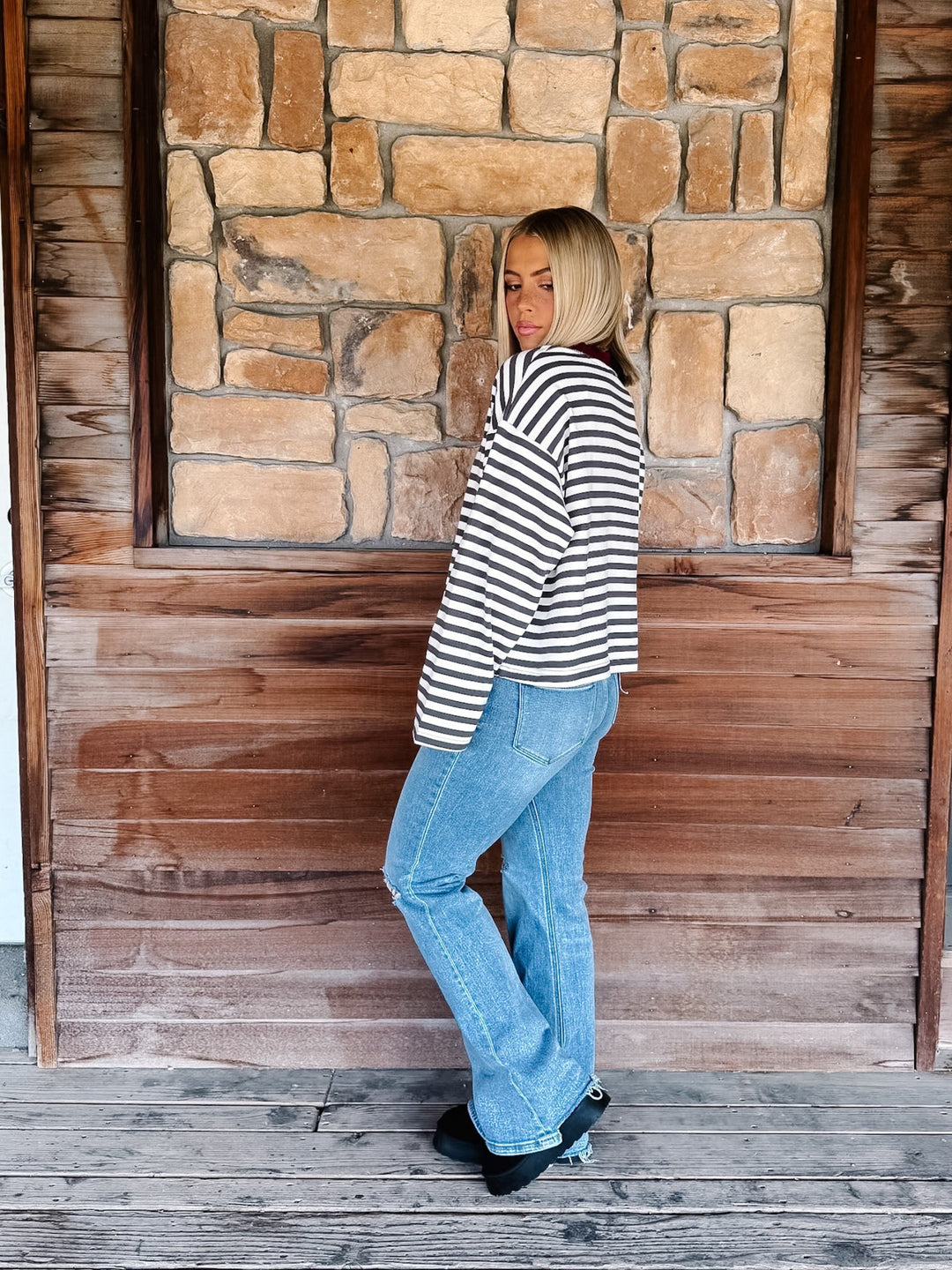 A woman with blonde hair wearing a charcoal and white horizontally striped long-sleeve top with maroon neckline detail and light blue jeans, standing in front of a rustic stone wall.