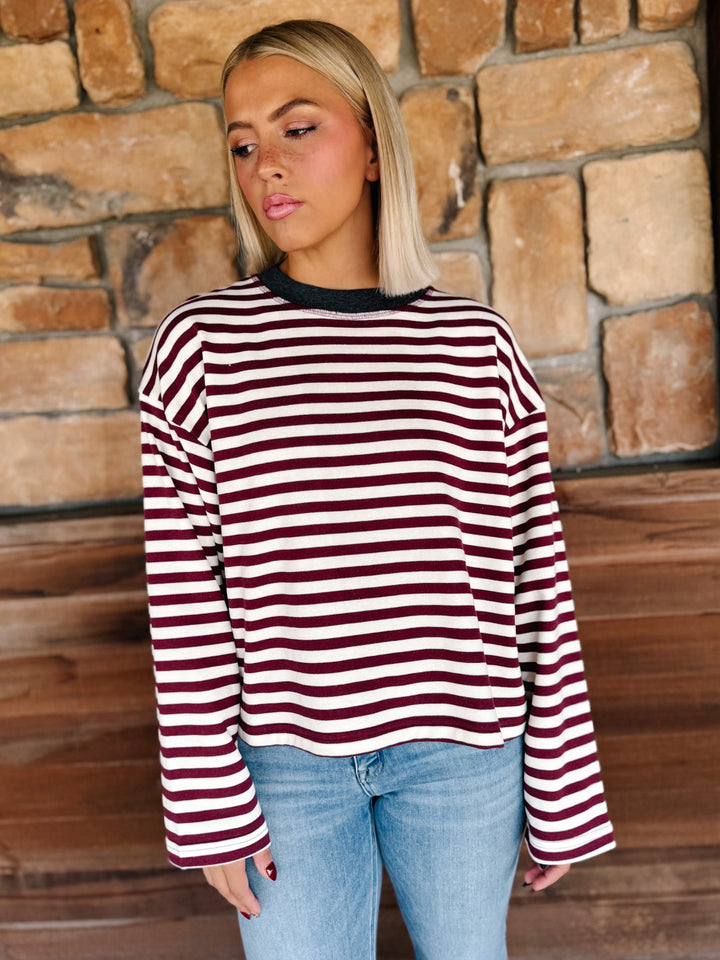 A woman with blonde hair wearing a long-sleeve maroon and cream striped sweater with a charcoal crew neck, paired with light blue jeans, standing in front of a rustic stone wall.