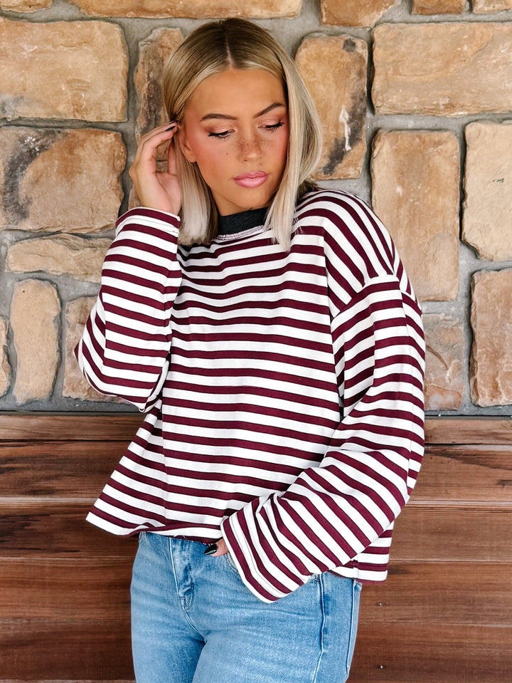 A woman with blonde hair wearing a long-sleeve maroon and cream striped sweater with a charcoal crew neck, paired with light blue jeans, standing in front of a rustic stone wall.