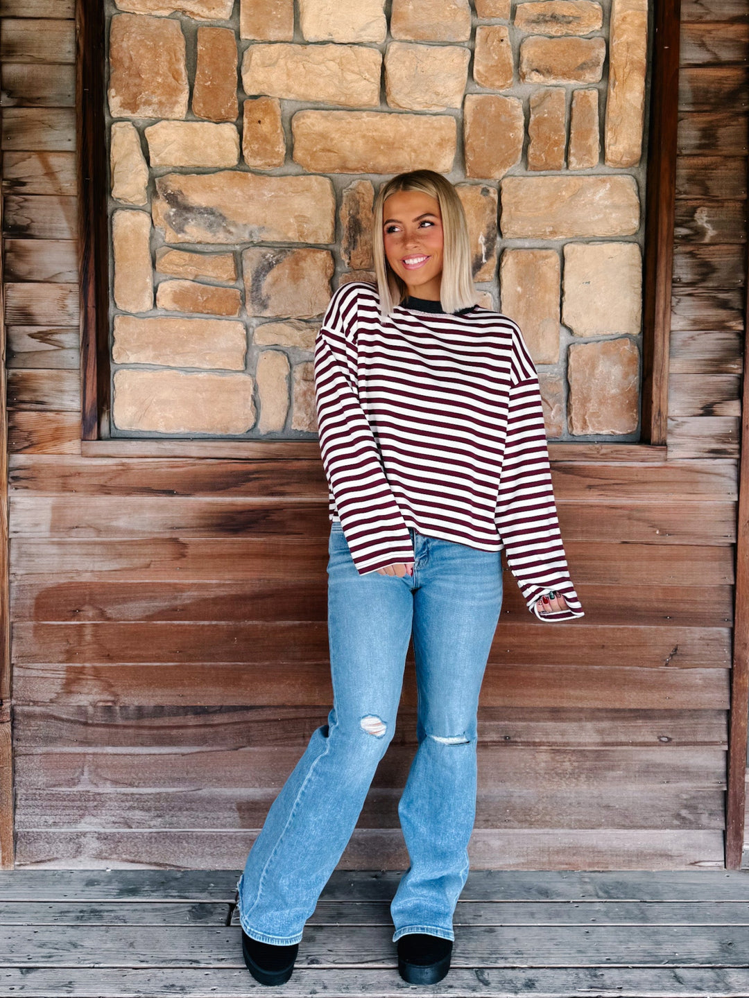A woman with blonde hair wearing a long-sleeve maroon and cream striped sweater with a charcoal crew neck, paired with light blue jeans, standing in front of a rustic stone wall.