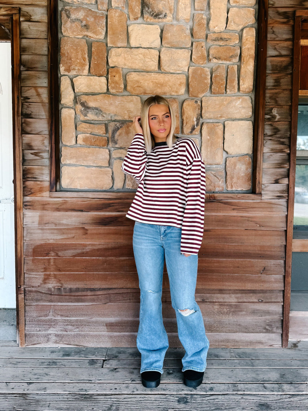 A woman with blonde hair wearing a long-sleeve maroon and cream striped sweater with a charcoal crew neck, paired with light blue jeans, standing in front of a rustic stone wall.