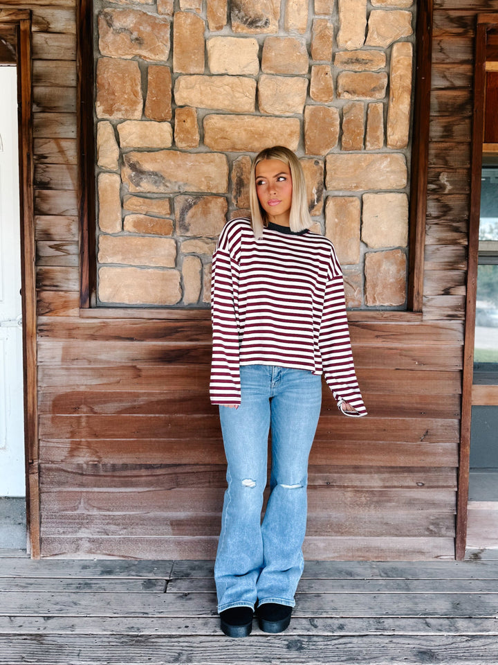 A woman with blonde hair wearing a long-sleeve maroon and cream striped sweater with a charcoal crew neck, paired with light blue jeans, standing in front of a rustic stone wall.