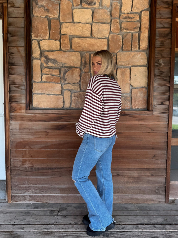 A woman with blonde hair wearing a long-sleeve maroon and cream striped sweater with a charcoal crew neck, paired with light blue jeans, standing in front of a rustic stone wall.