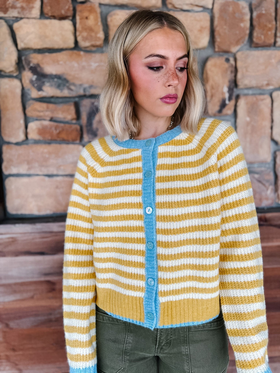 A woman wearing a yellow and white striped knit cardigan with a light blue button placket and collar, paired with olive green pants, standing in front of a rustic stone wall.