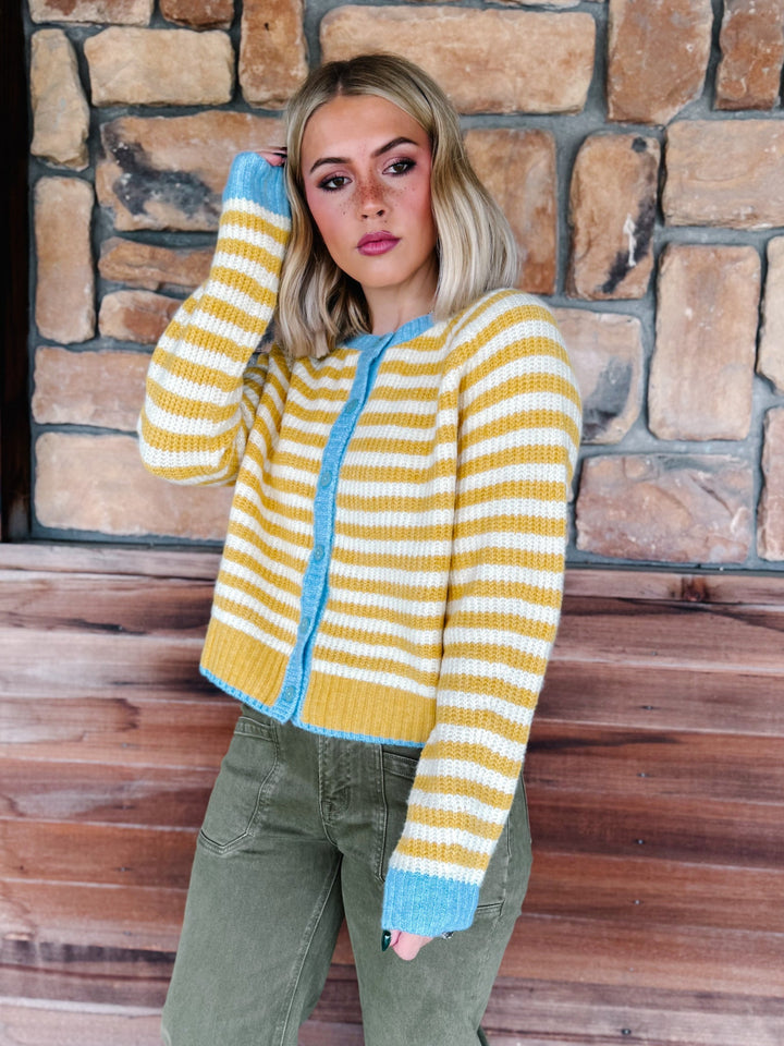 A woman wearing a yellow and white striped knit cardigan with light blue trim, paired with olive green pants, standing in front of a rustic stone wall and wooden porch.