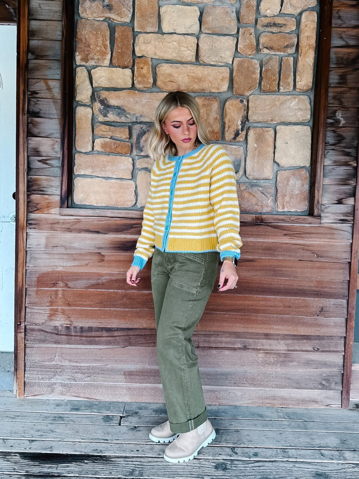 A woman stands on a rustic wooden porch in front of a stone wall, wearing a yellow and white striped knit cardigan with a bright blue zipper and trim, olive green pants, and beige boots.