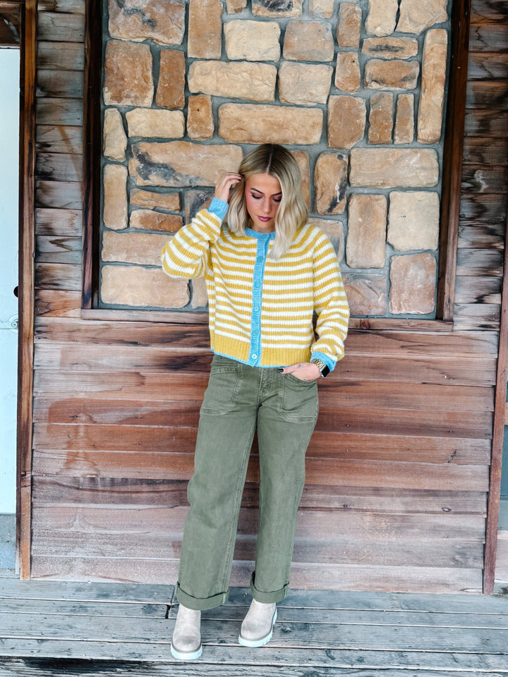 A woman stands on a wooden porch in front of a stone wall, wearing a yellow and white striped knit cardigan with a light blue zipper and trim, olive green pants, and beige boots.