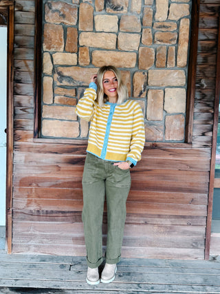 A woman stands on a wooden porch in front of a rustic stone and wood wall, wearing a yellow and white striped zip-up cardigan with light blue trim, olive green pants, and white sneakers.