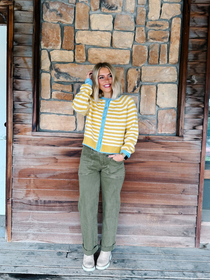 A woman stands on a wooden porch in front of a rustic stone and wood wall, wearing a yellow and white striped zip-up cardigan with light blue trim, olive green pants, and white sneakers.