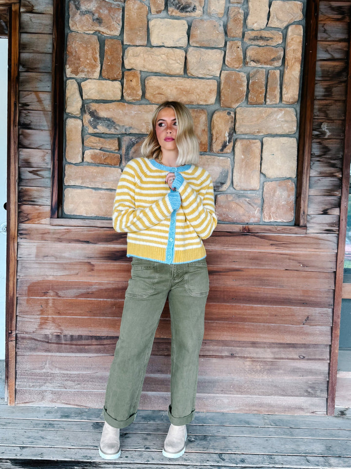 A woman stands on a wooden porch in front of a stone wall, wearing a yellow and white striped cardigan with a light blue collar, olive green pants, and white sneakers.