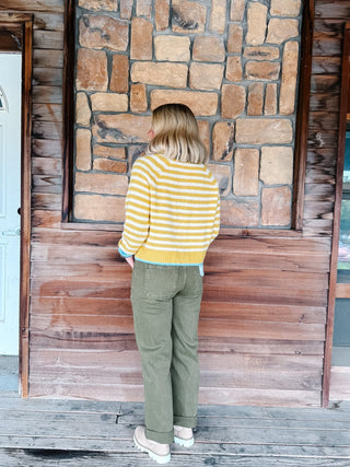 A woman stands with her back to the camera wearing a yellow and white striped knit sweater and olive green pants, standing on a wooden porch in front of a stone and wood wall.