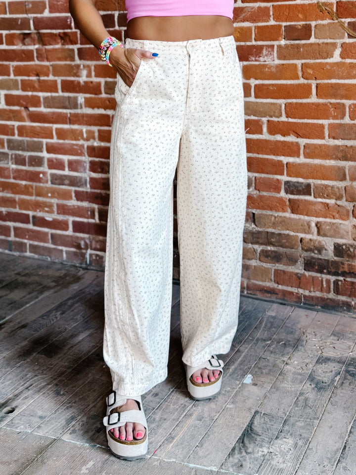 A person wearing loose, wide-leg cream pants with a small repeating floral print, standing against a red-brown brick wall and wooden floor.