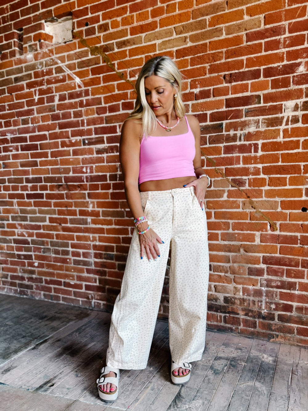 A woman posing in front of a brick wall wearing a bright pink ribbed tank top and cream-colored wide-leg patterned pants.