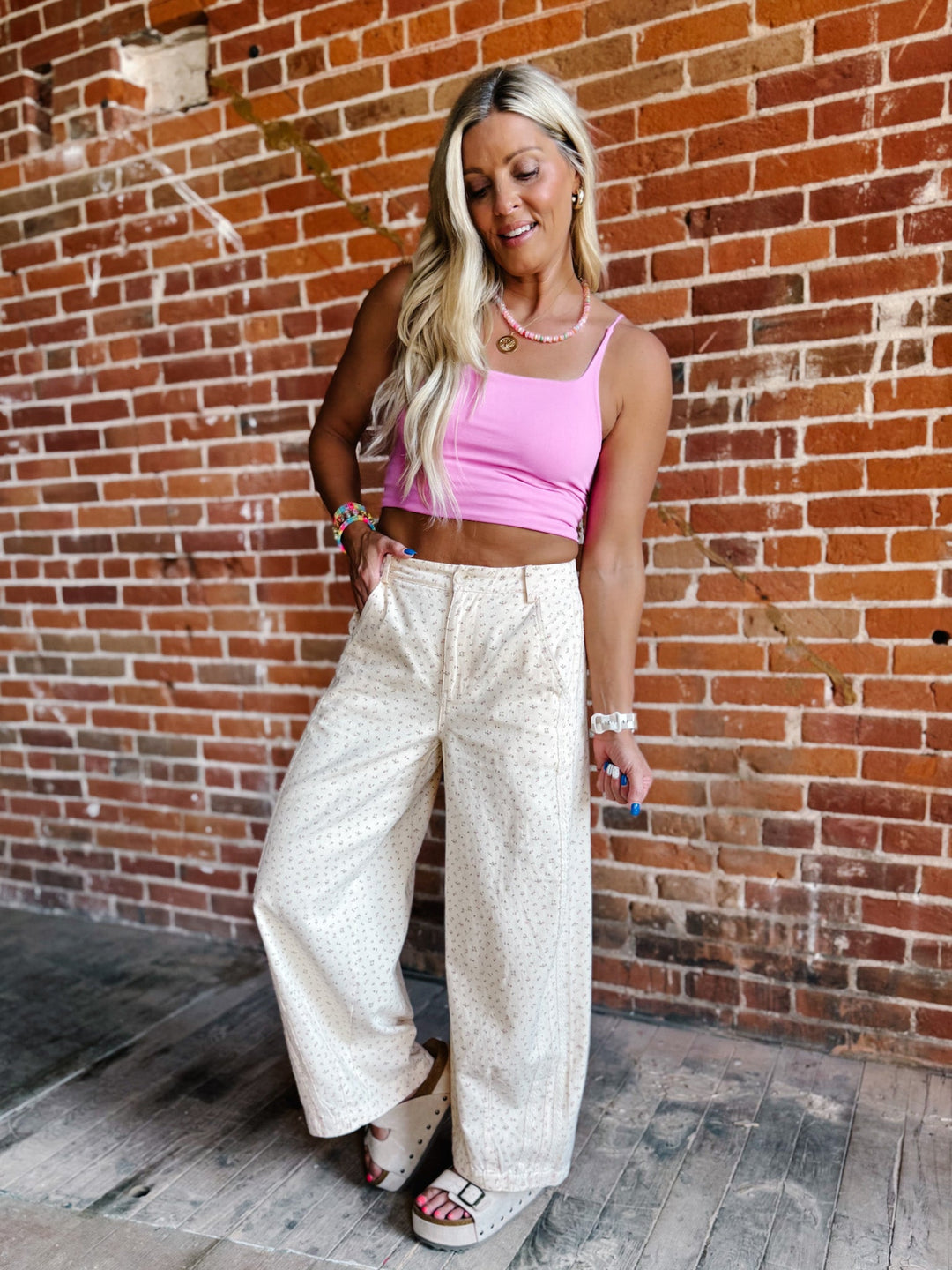 A woman posing in front of a red brick wall, wearing a bright pink crop top and cream-colored wide-leg patterned pants with white slide sandals.