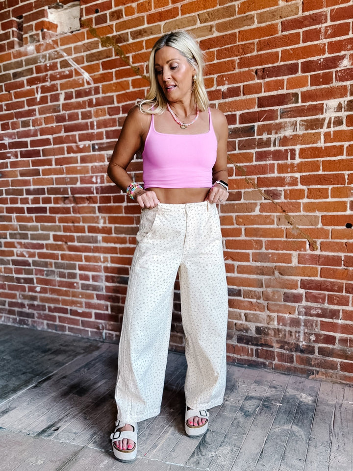 A woman stands against a red brick wall wearing a bright pink ribbed crop top and cream wide-leg pants with a small white pattern, paired with white slide sandals.