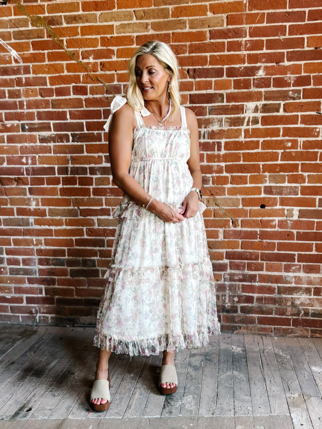 A woman wearing a floral chiffon dress with tie straps and a tiered silhouette, standing against a brick wall.