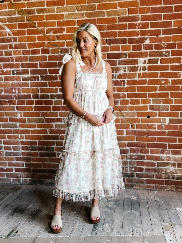 A woman wearing a floral chiffon dress with tie straps and a tiered silhouette, standing against a brick wall.