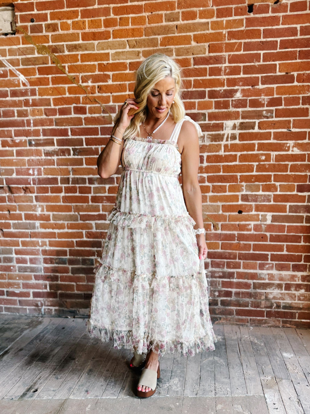 A woman wearing a floral chiffon dress with tie straps and a tiered silhouette, standing against a brick wall.