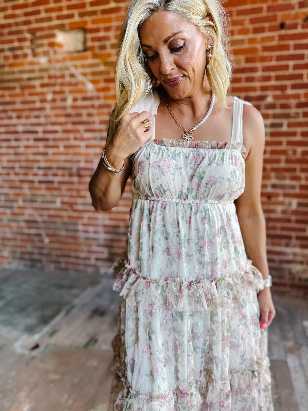 A woman wearing a floral chiffon dress with tie straps and a tiered silhouette, standing against a brick wall.