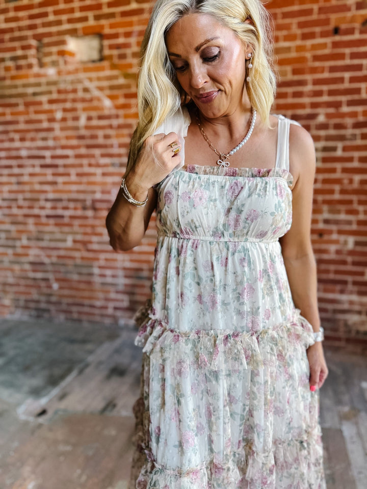 A woman wearing a floral chiffon dress with tie straps and a tiered silhouette, standing against a brick wall.