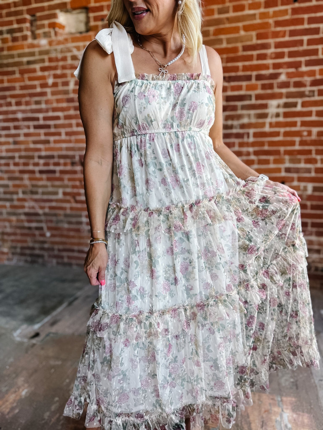 A woman wearing a floral chiffon dress with tie straps and a tiered silhouette, standing against a brick wall.
